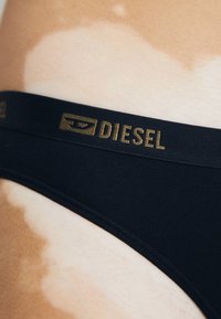 Svart bikini med en slät textur, med en gyllene "DIESEL"-logotyp på en elastisk midjeband. En kontrasterande hudton kan ses i närheten.