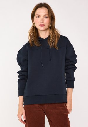 Frau mit heller Haut und roten Haaren trägt einen marineblauen Hoodie und braune Cordhosen und steht vor einem schlichten weißen Hintergrund.