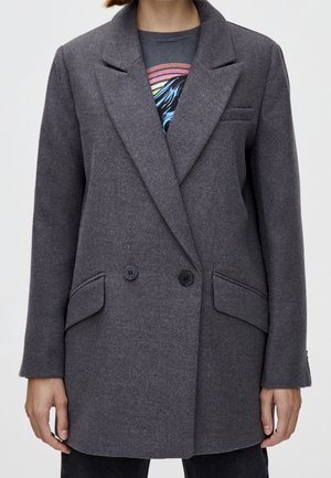 Personne portant un blazer gris croisé par-dessus un t-shirt noir avec un motif, debout les bras détendus le long du corps sur un fond clair.