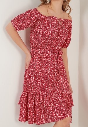 Femme portant une robe rouge à fleurs, épaules dénudées, manches bouffantes et ourlet à volants, posant avec une main sur la taille.
