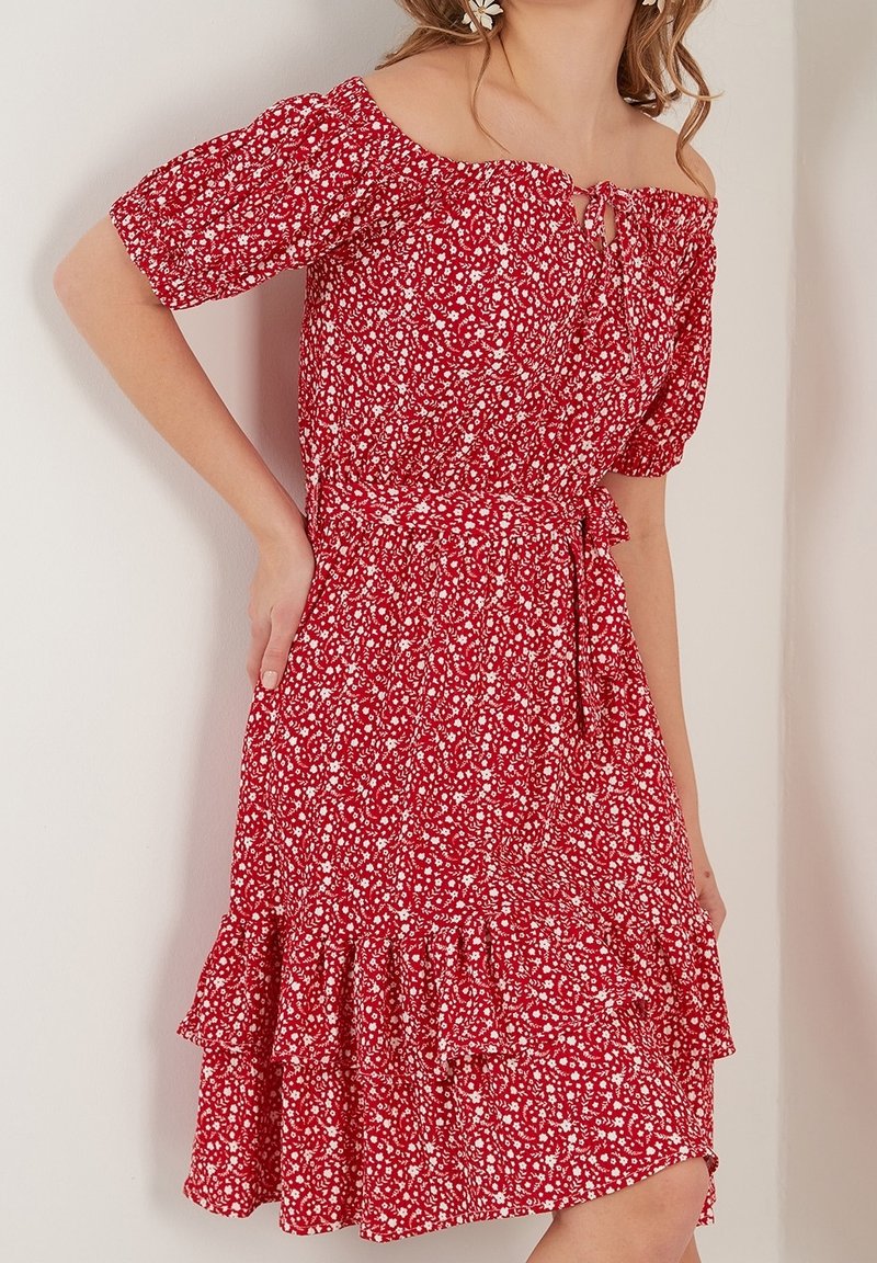 Femme portant une robe rouge à fleurs, épaules dénudées, manches bouffantes et ourlet à volants, posant avec une main sur la taille.