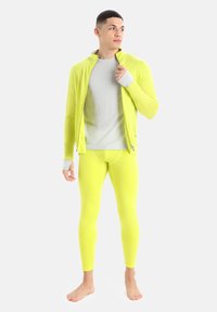 Neon žlutá sportovní bunda se zipem, spárovaná s odpovídajícími legínami; pod ní má šedé triko s dlouhým rukávem, hladká textura.