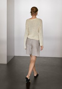 Kobieta odchodząca w beżowym swetrze, jasnoszarych szortach do kolan i czarnych klapkach na ciemnej podłodze.