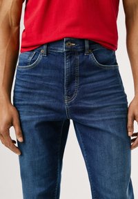 Temno modri kavbojki z oprijetim krojem, ki imajo pet žepov, rumene niti kot dodatek ter vidno zaponko na pasu.