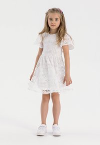 Robe en dentelle blanche avec des manches courtes à volants, des motifs complexes et une jupe évasée. Portée avec des baskets blanches, se tenant sur un fond neutre.