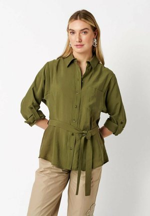 Frau trägt olivgrünes Hemd mit Knopfleiste und Stoffgürtel in der Taille, hochgekrempelte Ärmel, beigefarbene Hose mit Blumenstickerei.