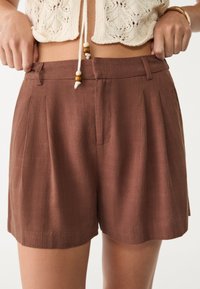 Brown pleated shorts with a textured fabric, featuring a mid-rise waistband and side pockets. Accented with a string detail at the waist.