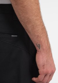 Pantalones cortos negros de Volcom con un bolsillo lateral, que presentan una etiqueta de logo visible. Primer plano de un brazo moreno con un tatuaje de rayo.