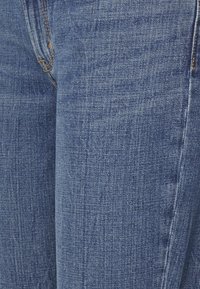 Os jeans de denim azul apresentam um corte justo, costura visível e uma ligeira textura típica do tecido. Nenhum padrão ou hardware distinto é notado.