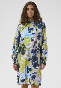 Žena s kovrčavom kosom koja nosi haljinu dugih rukava do koljena s šarenim apstraktnim tie-dye uzorkom u plavoj, zelenoj i crnoj boji.