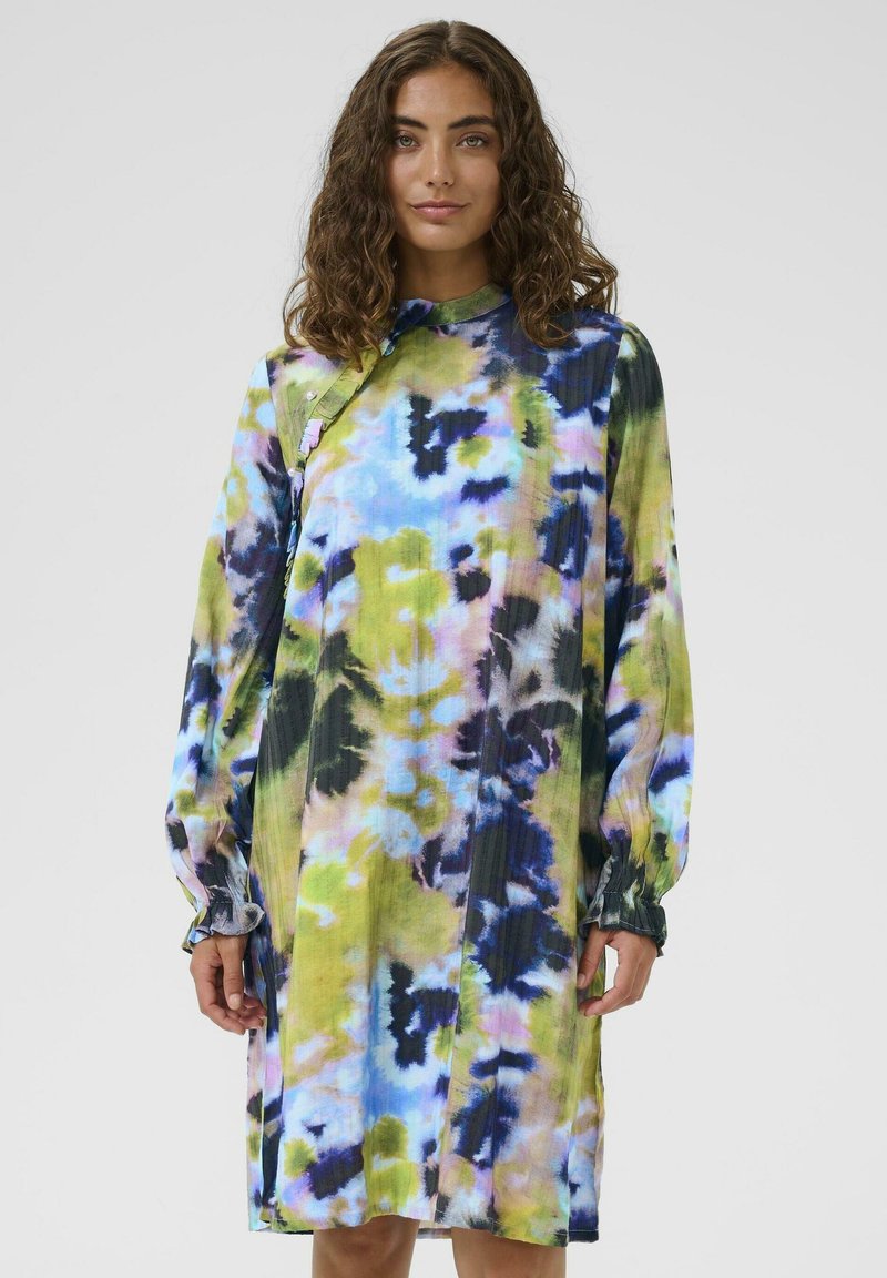 Žena s kovrčavom kosom koja nosi haljinu dugih rukava do koljena s šarenim apstraktnim tie-dye uzorkom u plavoj, zelenoj i crnoj boji.