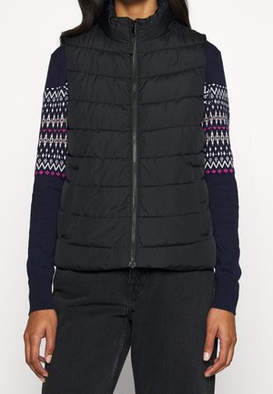 Vrouw draagt een zwart doorgestikt mouwloos vest over een marineblauwe gebreide trui met witte en roze details, gecombineerd met een zwarte broek.