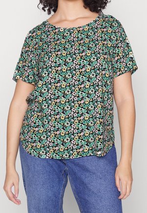 Femme portant un T-shirt noir ample avec un petit imprimé floral vert, rose et jaune, associé à un jean en denim bleu.