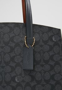 Sac cabas en cuir noir avec un motif texturé, doté d'une anse foncée et d'un détail en chaîne dorée sur une étiquette rectangulaire.
