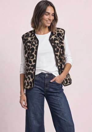 Femme portant un gilet en polaire imprimé léopard par-dessus une chemise blanche à manches longues et un jean bleu, souriant avec une main dans la poche.