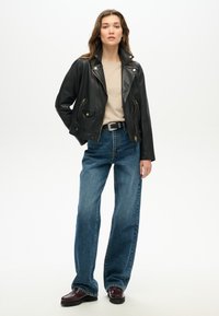 Veste en cuir noir de biker avec fermeture éclair à l'avant et fermetures à pression, portée sur une chemise beige ; associée à un jean bleu ample et des chaussures sombres.