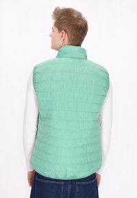 Minzgrüne Pufferweste mit hohem Kragen und horizontalen Nähten. Getragen über einem weißen Langarmshirt mit lockerer Passform.