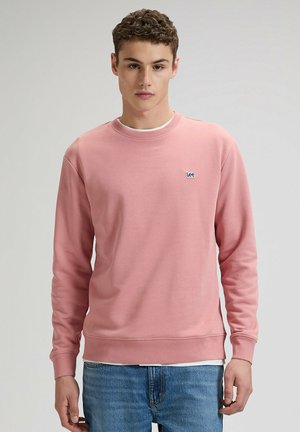 Jasnoróżowa bluza z miękkiego materiału z okrągłym dekoltem, długimi rękawami i małym niebieskim logo Lee na piersi. Do tego niebieskie dżinsy.