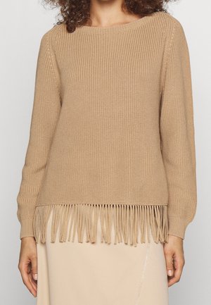Pull en tricot beige à manches longues avec un ourlet à franges porté par une personne aux cheveux bouclés, associé à une jupe beige clair.