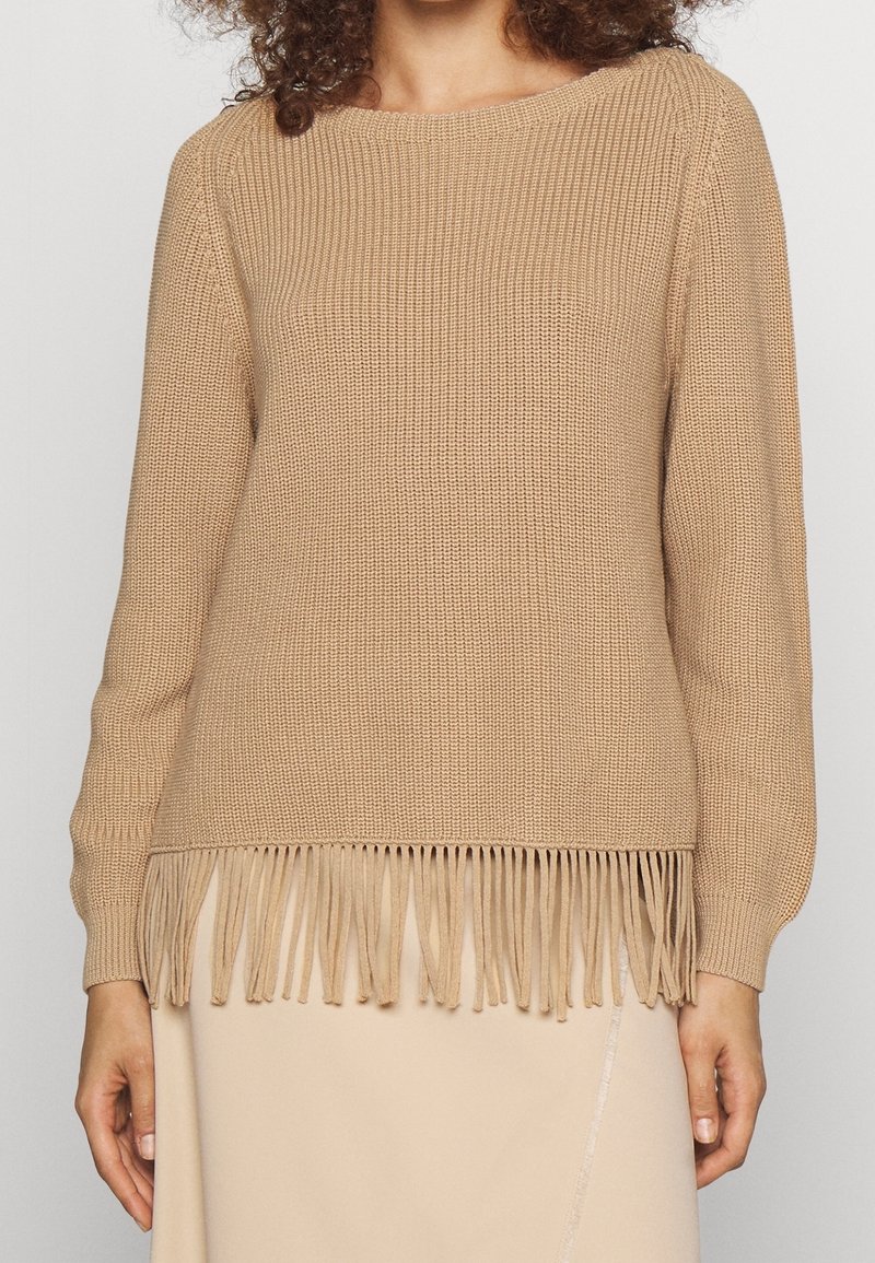 Pull en tricot beige à manches longues avec un ourlet à franges porté par une personne aux cheveux bouclés, associé à une jupe beige clair.