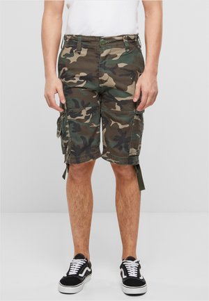Uomo che indossa pantaloncini cargo mimetici verdi e marroni, maglietta bianca e sneakers nere con lacci bianchi, in piedi su uno sfondo grigio uniforme.