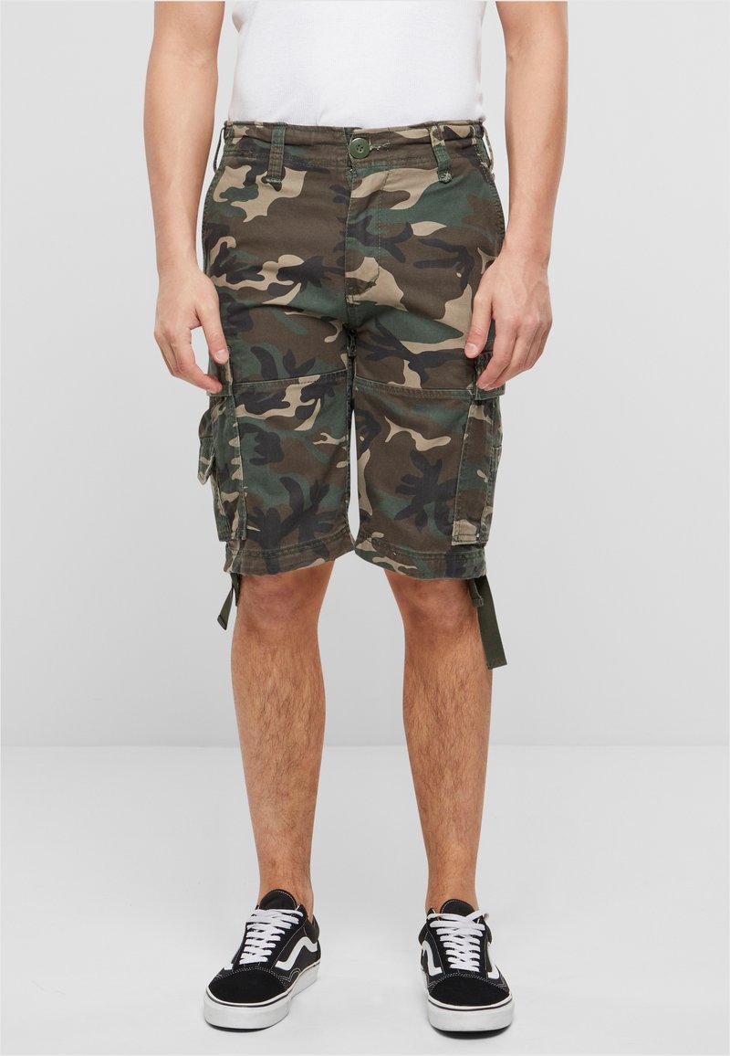 Homme portant un short cargo camouflage vert et marron, un t-shirt blanc et des baskets noires avec des lacets blancs, debout sur un fond gris uni.
