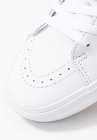 Vitt sportskor i slätt läder; har perforerade detaljer och kontrasterande sömmar längs ovansidan, med en texturerad gummisula.