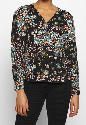 Vrouw draagt een zwarte blouse met lange mouwen met kleurrijke bloemenpatronen en zwarte broek met fijne strepen.