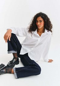 Bílá košile s dlouhými rukávy z lehké látky a s malým červeným akcentem na límci, doplněná tmavě modrými džíny s ohrnutými nohavicemi a černými botami.