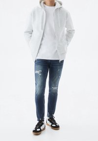 Jasnoszara bluza na zamek, biała koszulka, podniszczone niebieskie jeansy oraz czarno-białe sneakersy z gumowymi podeszwami. Codzienny strój o luźnym kroju.