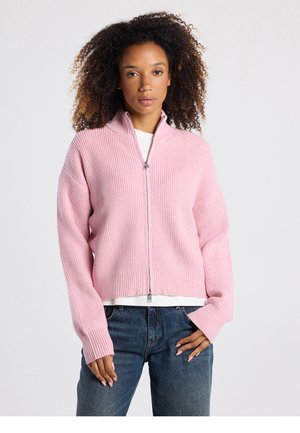 Roze vest met ritssluiting, geribbelde textuur, brede mouwen en een cropped ontwerp. Voorzien van een zilveren rits en gedragen over een witte top.