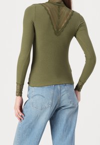 Olijfgroene langemouwtop met ingewikkelde kantdetails op de rug en manchetten, gestyled met losse blauwe spijkerbroeken.