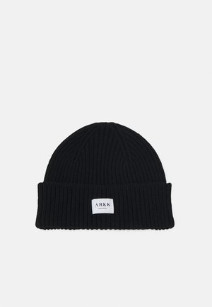 Gorro de malha preto com textura canelada e borda virada. Apresenta um pequeno rótulo branco com o nome da marca "ARKK" preso à parte da frente.