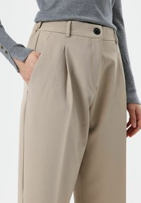 Beige trousers made of smooth fabric feature a high-waisted design with pleats, side pockets, and a single front button closure.