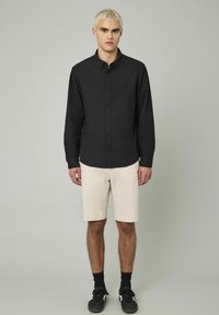 Chemise noire à manches longues avec boutons, en coton, col classique, portée avec un short beige et des baskets noires, debout sur un fond uni.