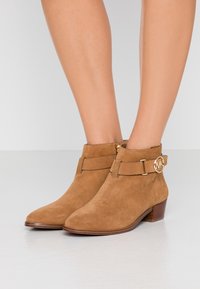 Voeten in tan suede enkelboots met lage gestapelde hakken en een gouden MK-logo gesp op de zijkanten tegen een witte achtergrond.