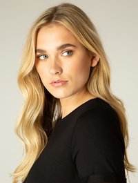 Blond haar dat in zachte golven valt; close-up van een gezicht met een lichte huid, gekleed in een zwart T-shirt met korte mouwen, met minimale make-up.