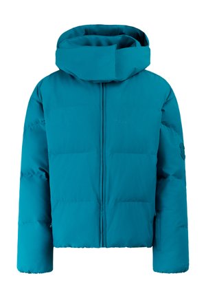 Jonge vrouw met zwarte muts, groene donzen jas, zwarte broek, handschoenen en laarzen, staand tegen een neutrale achtergrond.