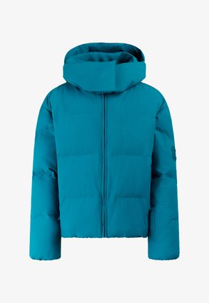 Jonge vrouw met zwarte muts, groene donzen jas, zwarte broek, handschoenen en laarzen, staand tegen een neutrale achtergrond.