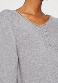 Harmaa neulepusero V-pääntiellä, jossa on pehmeä rakenne ja hillittyjen avoimien ommelkoristeiden yksityiskohdat saumoissa ja hartioissa.
