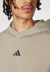 Grijs-groene hoodie van zacht materiaal, met een kangoeroezak en een zwart Adidas-logo op de borst. Ontspannen pasvorm met geribbelde accenten.