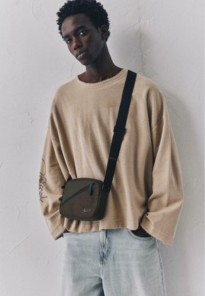Jeune homme portant un pull beige oversize et un jean clair, avec un petit sac bandoulière marron à sangle noire, devant un mur blanc texturé.