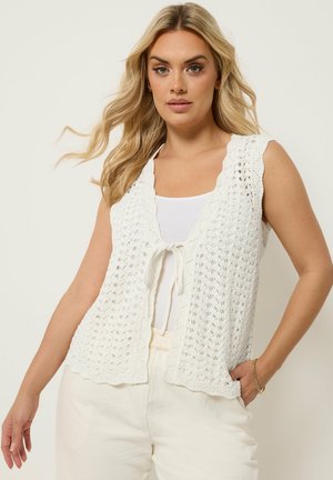Blondynka w białej, bezrękawnikowej kamizelce na szydełku, zawiązanej z przodu, na białym topie i białych spodniach, pozująca z jedną ręką w kieszeni.