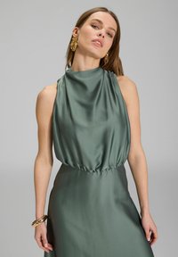 Robe sans manches en satin vert mat, fluide, avec un décolleté drapé et une taille froncée ; boucles d'oreilles dorées accrocheuses et bracelet texturé.