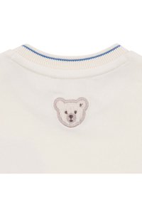 Tissu de couleur crème avec un col côtelé présentant une bande bleue et un petit visage d'ourson brodé centré sous l'encolure.