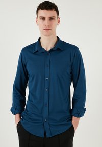 Sötétkék anyagú, hosszú ujjú ing, gallérral, sötét gombokkal ellátott gombolással és felgöngyölt ujjakkal.
