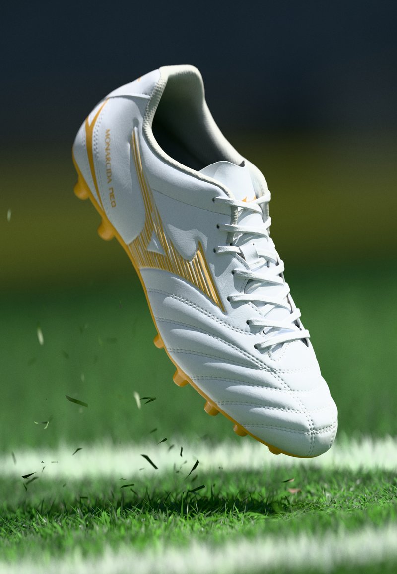 Scarpa da calcio Mizuno bianca e dorata con lacci, sospesa nell'aria sopra l'erba e una linea bianca del campo, sollevando fili d'erba.