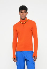 Oranžový pletený pulóver s vysokým golierom, ktorý obsahuje žlté a fialové akcenty. Dlhé rukávy s výrezmi, skombinované s modrými cargo nohavicami.