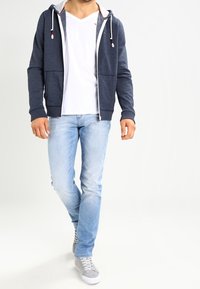 Marinblå zip-tröja med vit kant och röda detaljer, över en vit V-ringad t-shirt, parat med ljusblå jeans och grå sneakers.