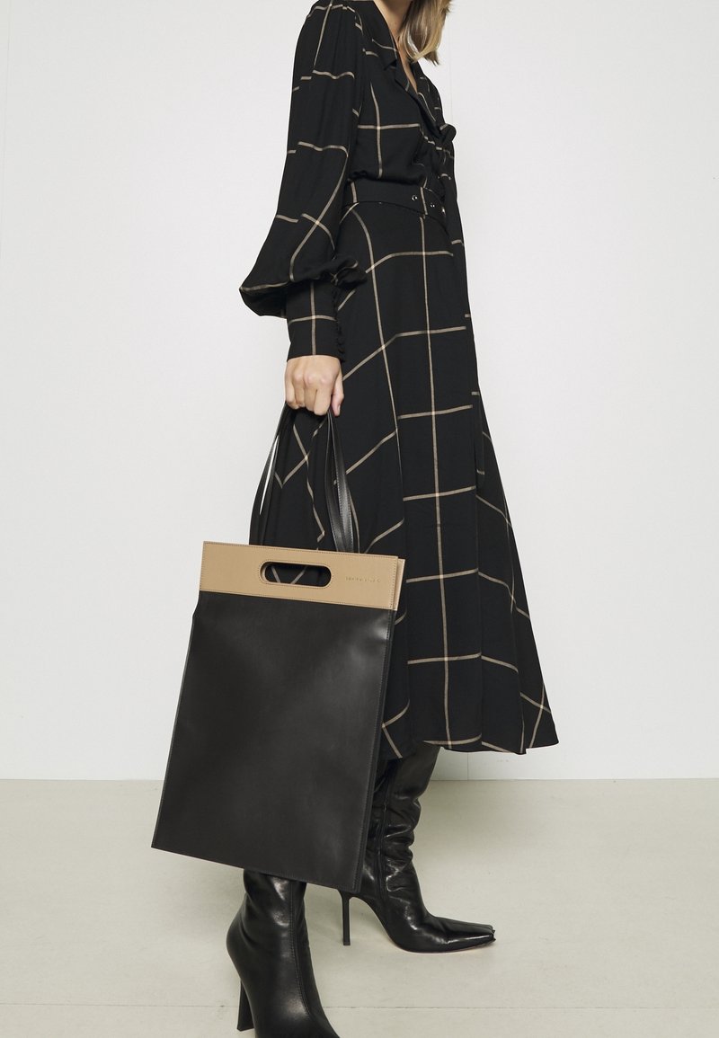 Femme en longue robe noire à carreaux et bottes à talons noires tenant un sac cabas en cuir noir et beige avec des doubles anses.